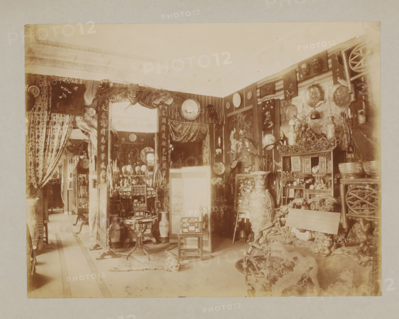 Interior, album page, photograph, This album page is part of a cover with 6 pages., collector's mark: 'Museum van Kunst-Nijverheid Haarlem', workshop stamp: 'J. Pörtzgen, Photograaf, Koningstraat 38, Haarlem', photographer: Johann Pörtzgen, c. 1890 - c. 1900, Johann Pörtzgen, photographer, paper, pasteboard, daylight collodion silver print, height 166 mm x width 220 mm, interior of the house, mirror, decorations (interior of the house), A sepia-toned interior filled with ornate decorative objects and furniture, including large vases, carved wooden chairs, a small round table, and shelves holding figurines and bowls; patterned curtains hang at openings and tapestries or framed panels cover walls, with circular plates and woven items displayed; a carved ornamental screen and a draped fabric panel appear near the center, while smaller stools and cabinets with intricate detailing occupy the floor space; various hanging ornaments, a cluster of sticks or poles, and a textured rug or animal h...