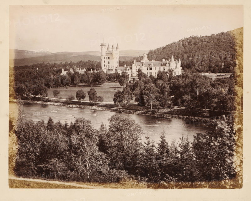 Balmoral, from the river, Balmoral Castle, album page, photograph, This page is part of an album., number: '315, JV', Aberdeenshire, c. 1875 - c. 1885, photographer, paper, albumen print, height 298 mm x width 236 mm, river, castle, A large pale stone building with multiple turrets and a taller central tower sits amid a landscaped lawn and clusters of trees, with a river flowing in the foreground and wooded hills rising behind the estate, all shown in a high-contrast monochrome photograph.