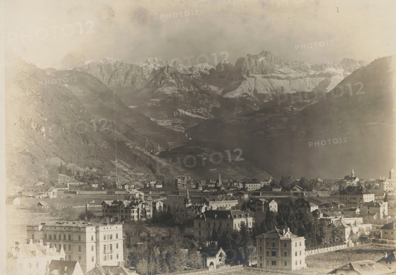 Landscape with mountains and a city, album page, photograph, This photo is part of an album., annotation: 'From Breitner's studio', 1880 - 1920, photographer, paper, pasteboard, gelatin silver print, developed gelatin silver print, height 129 mm x width 180 mm, Photo is loose in the front of the album., city-view, and landscape with man-made constructions, mountains, A panoramic view shows a town with clustered buildings and multi-story houses in the foreground, some with pitched roofs and decorative facades, interspersed with trees and fenced yards; further back a denser area of rooftops and church spires rises along a gentle slope leading into terraced fields and vineyards on the lower mountain slopes; beyond those the terrain steepens into dark, forested hills that give way to a dramatic range of jagged, snow-capped mountains under a hazy sky, with soft gradients of shadow and light across the valleys.