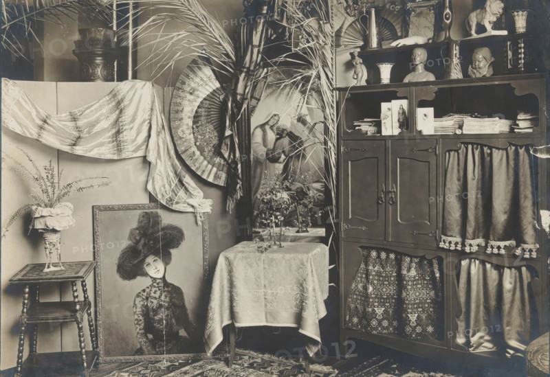Interior with a cupboard, sculptures and a painting, album page, photograph, This photo is part of an album., annotation: 'From Breitner's studio', 1880 - 1920, photographer, paper, pasteboard, gelatin silver print, developed gelatin silver print, height 129 mm x width 180 mm, Photo is loose at the front of the album., sculpture, picture, painting, interior of the house, furniture to put things on, furniture for storage, A room corner filled with decorative objects and furnishings, including a wooden cabinet with shelves displaying vases, busts, a small figurine, folded textiles, and stacked papers, curtains with fringe hanging from some openings, and patterned fabric visible behind lower openings; a table covered with a patterned cloth supporting a small plant and a tray with glassware; a large framed portrait of a woman wearing a hat leaning against the wall near a small turned-leg side table that holds a pedestal planter with a fern and a ruffled fabric cover; a draped silk or satin...