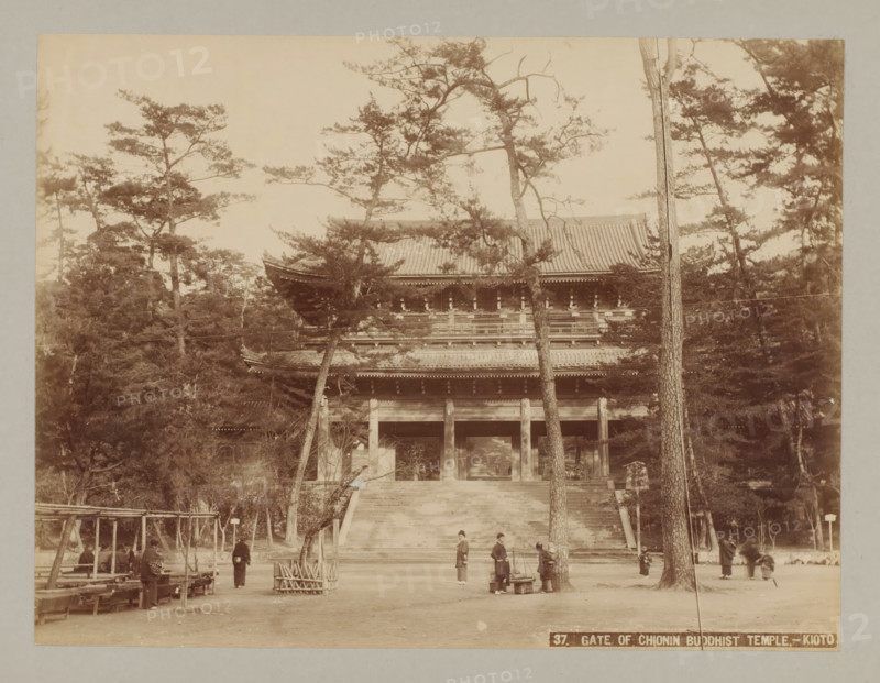 Gate of Chion-in Temple, Kyoto, 37. Gate of Chionin Buddhist Temple. - Kyoto, photograph, number: '6.', c. 1895 - c. 1915, photographer, Kinki, paper, pasteboard, albumen print, height 207 mm x width 273 mm, height 243 mm x width 329 mm, A wide sepia photograph shows a large traditional wooden gate with a tiled roof partially obscured by tall trees with thin trunks and spreading branches; wide stone steps lead up to the gate and a few people in traditional clothing stand or walk near the base of the steps, with some gathered around a carrying pole and baskets; to the left are simple wooden benches and a pergola-like wooden structure, while the foreground is an open dirt area with scattered small objects and tree stumps.