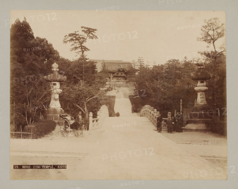 75. Nishi-Otni Temple.-Kioto, Nishi-Otani Temple, Kyoto, photograph, number: '7.', c. 1895 - c. 1915, photographer, Kinki, paper, pasteboard, photo paper on cardboard, height 210 mm x width 270 mm, height 243 mm x width 329 mm, A wide view of a stone bridge leading toward a distant temple gate framed by tall trees on both sides, with ornate stone lanterns atop pedestals flanking the bridge entrance, a rickshaw parked near the left lantern with a man standing beside it, a group of people standing near the right lantern, stone railings lining the bridge, trees and foliage surrounding the pathway, and a roofed temple structure visible beyond the top of the steps.