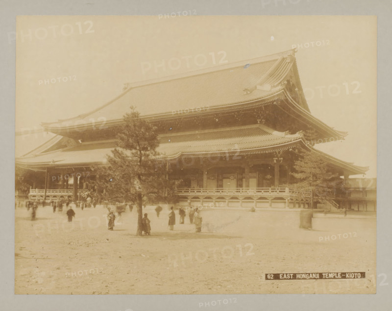 62 East Honganji Temple-Kioto, East side of Nishi Honganji Temple, Kyoto, photograph, number: '8.', c. 1895 - c. 1915, photographer, Kinki, paper, pasteboard, albumen print, albumendruk, height 209 mm x width 272 mm, height 243 mm x width 329 mm, A sepia-toned photograph showing a large traditional wooden temple building with a multi-tiered sweeping roof and ornate eaves, a raised veranda with railings, columns, and carved details, a tree in the foreground partially obscuring the temple, and a flat open ground in front where several people in period clothing stand or walk.