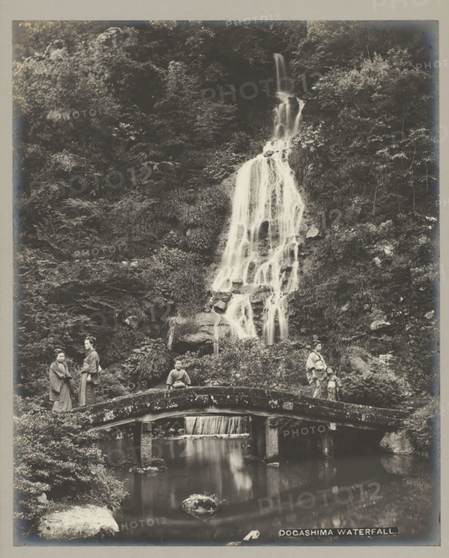 Dogashima waterfall., Dogashima waterfall, Kanagawa-Ken, photograph, number: '19.', c. 1895 - c. 1915, photographer, Kanto, paper, pasteboard, gelatin silver print, developing gelatin silver print, height 283 mm x width 229 mm, height 329 mm x width 243 mm, A tall, multi-tiered waterfall cascades down a rocky, vegetated hillside into a pool below, with water flowing over rocks and through grassy vegetation; a stone arched bridge spans the pool in the foreground, its surface mottled and textured, and beneath the bridge a smaller fall feeds the reflective water; on the bridge and near it stand several people wearing traditional garments, including adults and children, positioned at different points along the bridge and banks, with one person seated on the bridge; dense foliage and trees cover the surrounding slopes, and rocks and plants frame the scene on both sides.