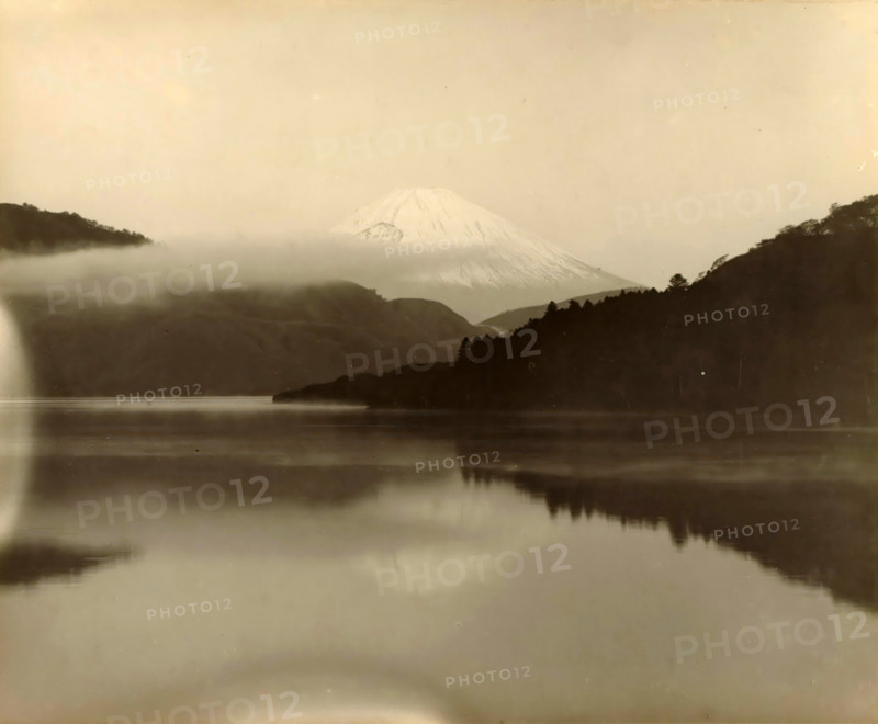 Fuji seen from a distance, Japan, photograph, annotation: '23. Fuji', c. 1895 - c. 1915, photographer, paper, pasteboard, gelatin silver print, developing gelatin silver print, height 229 mm x width 281 mm, height 243 mm x width 329 mm, A wide, misty landscape with a snow-capped conical mountain in the distance partially veiled by low clouds, darker forested hills on either side framing the mountain, a calm reflective body of water in the foreground mirroring the hills and sky, and a soft, pale sky above.