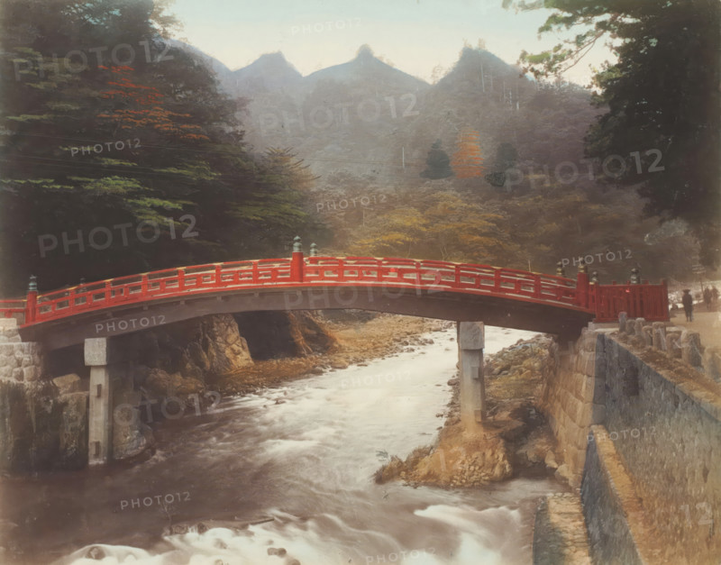 Sacred bridge, Japan, photograph, annotation: '27. Sacred bridge.', c. 1895 - c. 1915, photographer, paper, pasteboard, salted paper print, Salt print, colored., height 208 mm x width 267 mm, height 243 mm x width 329 mm, A red arched wooden bridge spans a flowing river, supported by stone and concrete piers, with carved railings and decorative finials along its length; rocky banks and a stone retaining wall border the river, and a pathway runs beside the wall. Dense trees and foliage frame both sides of the scene, with a mix of dark green and autumnal leaves visible, and distant misty mountains rise in the background under a pale sky. Two small human figures stand near the far end of the bridge, partially obscured, and scattered rocks and shallow rapids appear in the river beneath the bridge.