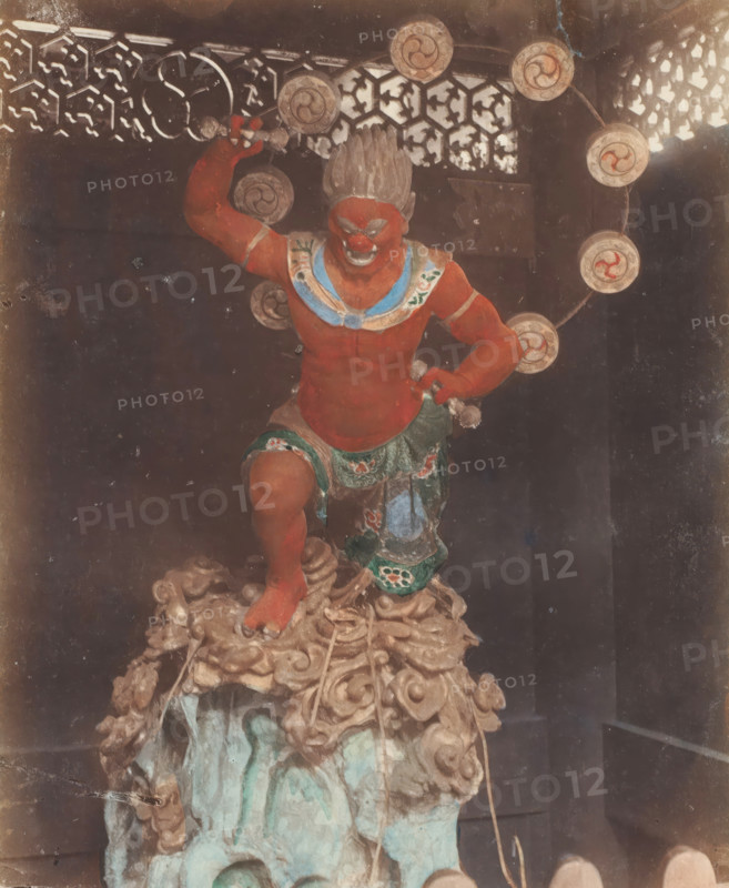 B 85 The Thunder God of Buddest Temple Nikko, God of Thunder at Buddhist temple, Nikko, photograph, number: '31.', c. 1895 - c. 1915, photographer, Kanto, paper, pasteboard, salted paper print, Salt print, colored., height 253 mm x width 201 mm, height 329 mm x width 243 mm, A painted statue of a muscular red-skinned figure stands on a carved base that resembles swirling clouds and rock, the figure posed with one leg raised and one arm bent upward holding a small object, wearing a decorative skirt with green and patterned details and a blue and white collar, the face showing a fierce expression with prominent brows and a wide mouth and hair styled upward, a circular halo of disks with spiral motifs arches behind the figure, an ornate lattice screen with geometric cutouts is visible in the upper background, and the image shows signs of age with scratches and discoloration.