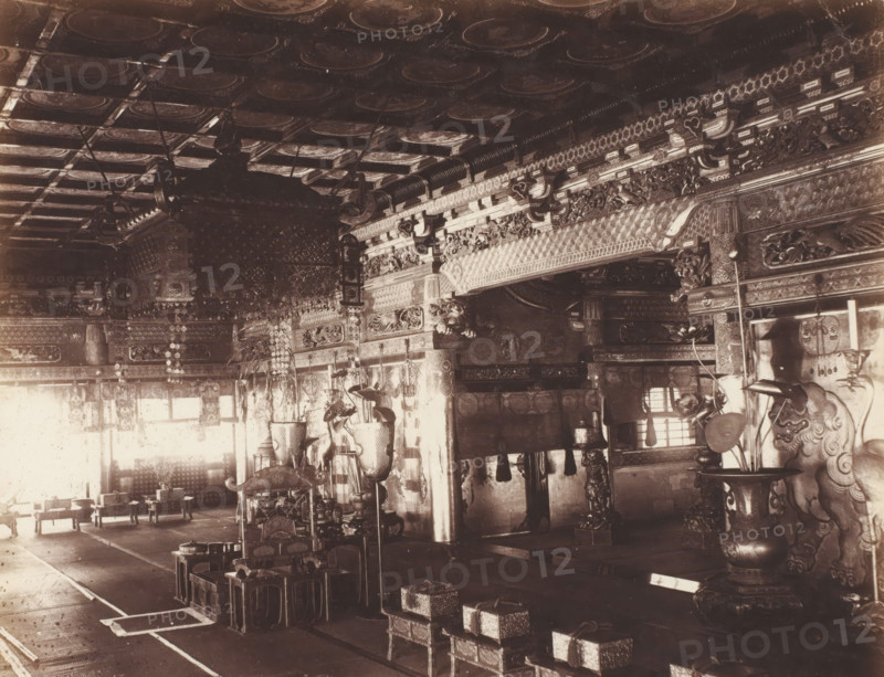 B 3 A front hall of Buddest Temple Nikko., Hall in the front of Buddhist temple, Nikko, photograph, number: '3.3.', c. 1895 - c. 1915, photographer, Kanto, paper, pasteboard, salted paper print, Zoutdruk, height 210 mm x width 265 mm, height 243 mm x width 329 mm, An ornate interior space with a decorated coffered ceiling and carved beams, large hanging lanterns and decorative cords suspended from above, intricately carved wooden pillars and beams with repeating geometric and floral motifs along the upper walls, rows of low wooden stools and small ornate boxed platforms arranged on the floor, tall ornamental vases and stands with sculpted details positioned near the front, carved guardian figures flanking the sides with curled manes and stylized faces, hanging decorative garlands and plaques along the walls, patterned floor coverings and runners defining walkways, and light coming through windows that casts shadows across the furnishings.