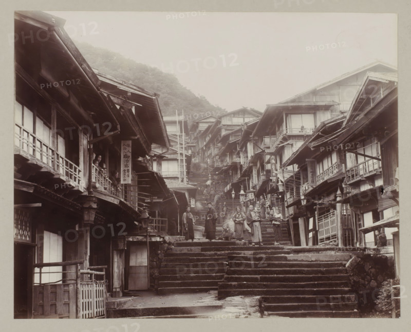 Street in Ikaho with steps, photograph, annotation: '42. Ikao.', Ikaho, c. 1895 - c. 1915, photographer, Kanto, paper, pasteboard, daylight collodion silver print, daylight collodion silver print on photo paper on cardboard, height 201 mm x width 260 mm, height 243 mm x width 329 mm, A narrow stone stairway ascends between closely packed wooden buildings with overhanging eaves and balconies, the façades showing exposed beams and lattice windows; people in traditional clothing stand and walk on the steps and on the balconies, some holding children, others gathered in small groups; wooden signboards hang from several buildings but their content is not described; the street slopes upward toward a misty hillside with trees visible in the background, and lanterns and railings line portions of the stairs and building fronts.