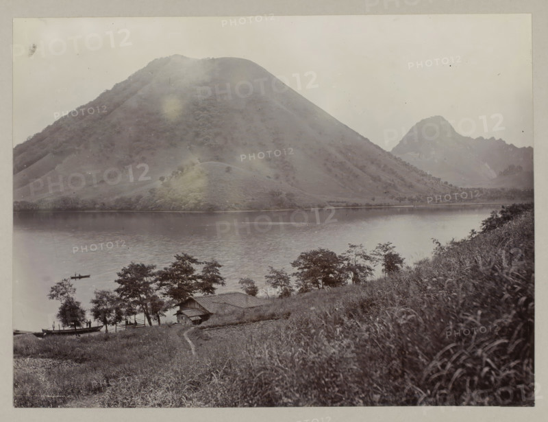 More from Haruna, photograph, annotation: '44. Haruna lake.', Haruna, c. 1895 - c. 1915, photographer, Kanto, paper, pasteboard, daylight collodion silver print, daylight collodion silver print on photo paper on cardboard, height 210 mm x width 280 mm, height 243 mm x width 329 mm, A wide lake fills the middle ground, its surface smooth and reflective, bordered by a shoreline with scattered trees and grassy slopes; a large rounded mountain rises steeply from the far shore, covered in textured vegetation, and a smaller jagged peak appears further back to the right; in the foreground a hillside with tall grasses slopes down toward a rustic wooden building with a pitched roof nestled among trees near the water, and a narrow footpath leads down the slope; small boats float on the lake, one with a person standing, and leafy trees frame the lower edge of the scene.