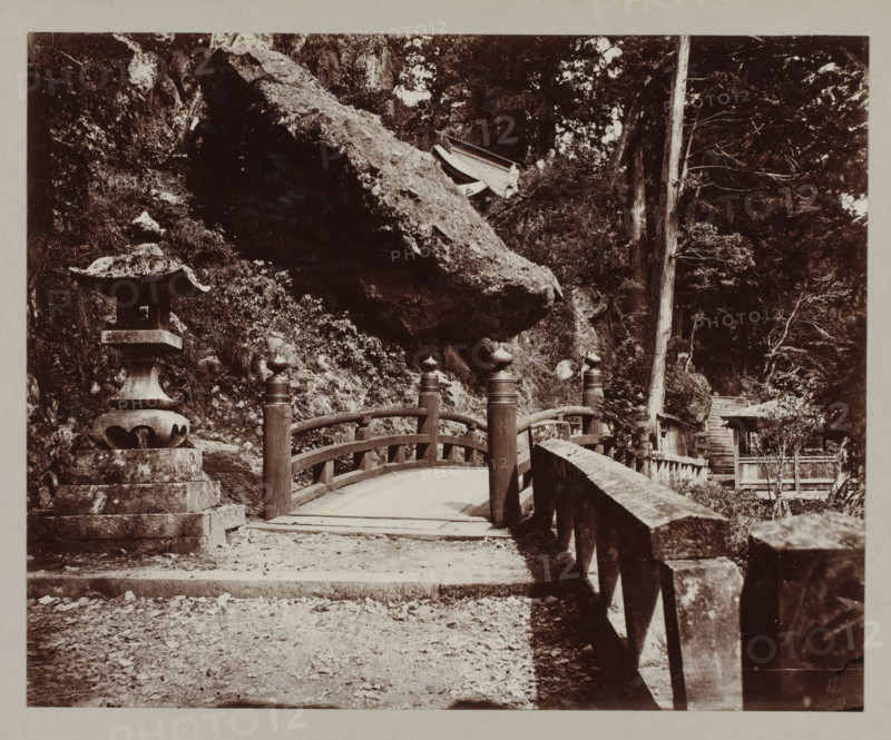 Entrance of the Haruna temple, photograph, annotation: '46. Entrance to Haruna temple.', Haruna, c. 1895 - c. 1915, photographer, Kanto, paper, pasteboard, daylight collodion silver print, daylight collodion silver print on photo paper on cardboard, height 217 mm x width 268 mm, height 243 mm x width 329 mm, A stone lantern stands on a stepped base to the left of a gently arched wooden bridge with rounded posts topped by bulbous finials, the bridge leading toward a rocky hillside and dense trees; a large overhanging rock looms above the scene, and a worn stone railing runs along the right side of the path, with scattered pebbles and leaves on the ground and a set of stone steps and small structures partially visible among the trees in the background.