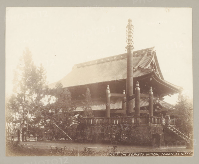 KS 3 The Sorinto Buddhi Temple at Nikko., Temple with Sorinto pillar, Nikko, photograph, c. 1895 - c. 1915, photographer, Kanto, paper, pasteboard, salted paper print, Zoutdruk., height 210 mm x width 265 mm, height 243 mm x width 329 mm, A sepia photograph showing a large traditional temple building with a steep tiled roof and ornate curved eaves, a tall decorative pillar rising in front of the roof, a stone platform with wooden railings and stout posts visible at the base of the structure, trees and foliage partially obscuring the lower sides of the building, and a set of stone steps leading up to the platform.