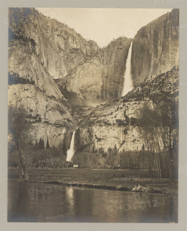 Yosemite waterfall, photograph, Bottom right reads: 10109., annotation: '2. Yosemete-fall.', California, c. 1895 - c. 1915, photographer, North America, paper, pasteboard, gelatin silver print, developing gelatin silver print, height 238 mm x width 189 mm, height 329 mm x width 243 mm, A sepia-toned landscape photograph showing a tall, steep rock face with two prominent waterfalls cascading down different sections of the cliff; the upper waterfall falls from near the top of the rock into a misty basin below while the lower waterfall continues down to a tree-lined valley; rocky slopes and scattered trees cover the cliff surfaces, with clusters of trees at the base and leafless trees along the right foreground; a reflective river runs across the bottom of the image with rippled reflections and a small grassy bank; a small building with a pitched roof sits near the treeline, partially obscured by trees.