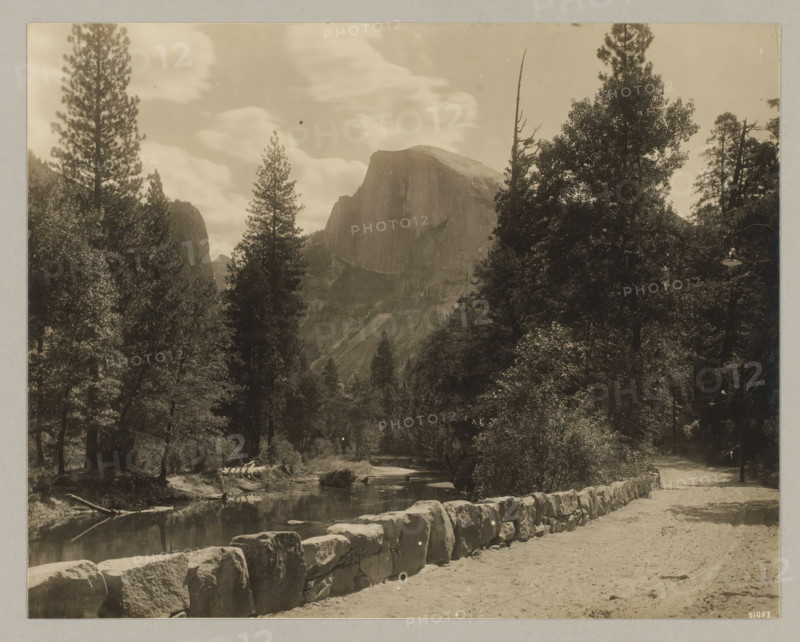 Yosemite Valley, photograph, Bottom right reads: 51083., annotation: '3. Yosemete Valley', California, c. 1895 - c. 1915, photographer, North America, paper, pasteboard, gelatin silver print, developing gelatin silver print, height 189 mm x width 239 mm, height 243 mm x width 329 mm, A sepia-toned landscape photograph showing a rocky mountain with a rounded top framed by tall conifer trees and dense foliage, a calm river running through the foreground with reflections of nearby trees, a low stone wall lining a dirt path beside the river, scattered large boulders and logs near the water, and a lamp post partially visible among the trees along the path.