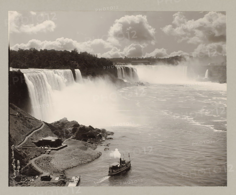 Niagara Falls, United States, Canada, photograph, a boat is sailing in the middle at the bottom., annotation: '7. Niagara Fall seen from the Canadian side', photographer: Landreth, 1910, North America, paper, pasteboard, gelatin silver print, height 193 mm x width 241 mm, height 243 mm x width 329 mm, A wide view of a large waterfall with water cascading over a cliff into a misty plunge pool, a rocky shoreline with a winding path and a small building near the edge, a steamboat moving on the river leaving a plume of smoke, a tree-covered ridge above the falls, low clouds filling the sky, and a mist veil rising where the water meets the river.