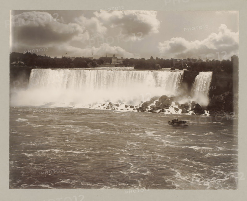Niagara waterfall, United States, Canada, photograph, Niagara waterfall seen from the front, on the right a boat sails., number: '8.', Niagara Falls (North America), c. 1895 - c. 1915, photographer, North America, paper, pasteboard, gelatin silver print, developing gelatin silver print, height 182 mm x width 239 mm, height 243 mm x width 329 mm, A wide view of a large waterfall with water cascading over a cliff into a misty, rocky plunge below, a river with rippling, foamy water in the foreground, a small boat on the river near the waterfall, rocky outcrops and spray at the waterfall base, a tree-lined shoreline and buildings visible on the far bank above the falls, and a sky filled with large, billowing clouds.