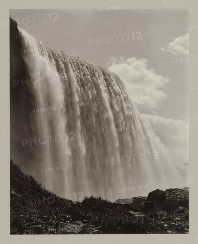 Niagara waterfall seen from below, United States, Canada, photograph, number: '9.', Niagara Falls (North America), c. 1895 - c. 1915, photographer, North America, paper, pasteboard, gelatin silver print, developing gelatin silver print, height 239 mm x width 189 mm, height 329 mm x width 243 mm, A large curtain of water cascades over a cliff, filling the left and central parts of the image with rushing, misty flows; the water appears textured with streams and softer, blurred areas where it falls. A vertical dark rock face is visible at the top left where the water begins to spill over. Clouds occupy the upper right portion of the scene, appearing soft and diffuse against a pale sky. In the lower foreground there is rocky ground with patches of grass and low vegetation, and a few larger rocks along the bottom edge. The overall image is monochromatic with high contrast between the bright falling water and the darker rocks and vegetation.