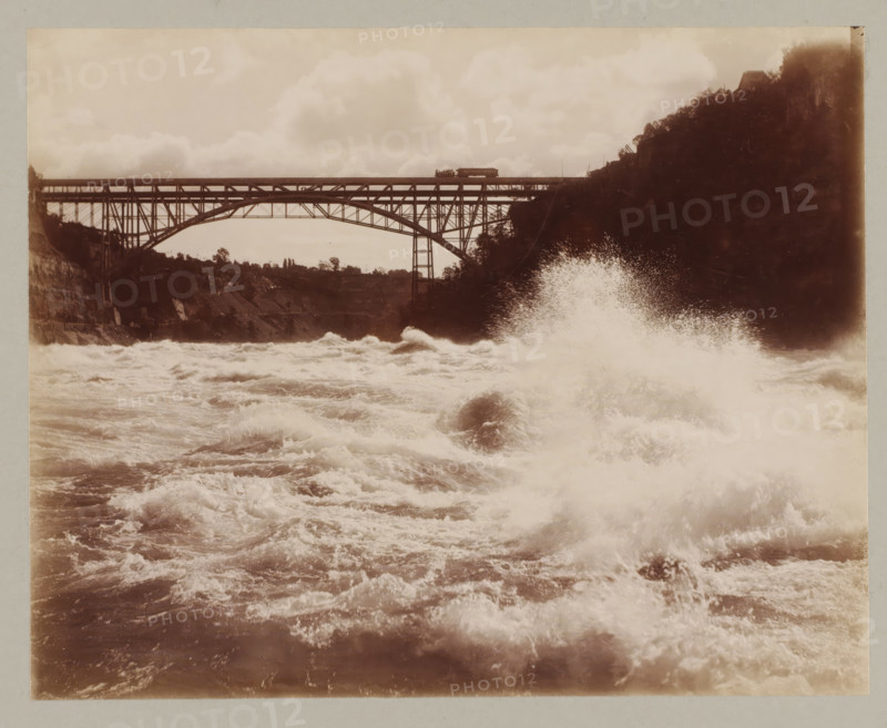 Bridge between America and Canada, United States, Canada, photograph, Bridge between America and Canada seen from the water., annotation: '10. Bridge between Canada & America', Niagara Falls (North America), c. 1895 - c. 1915, photographer, North America, paper, pasteboard, albumen print, height 193 mm x width 243 mm, height 243 mm x width 329 mm, bridge, A wide sepia-toned photograph showing turbulent water in the foreground with frothy waves and spray rising, a large spray plume toward the right side, and a steel arch bridge spanning the scene in the midground with visible trusses and supports; a small rail car sits on the bridge, cliffs and tree-covered slopes flank both sides of the river, and cloudy sky fills the upper portion of the image.