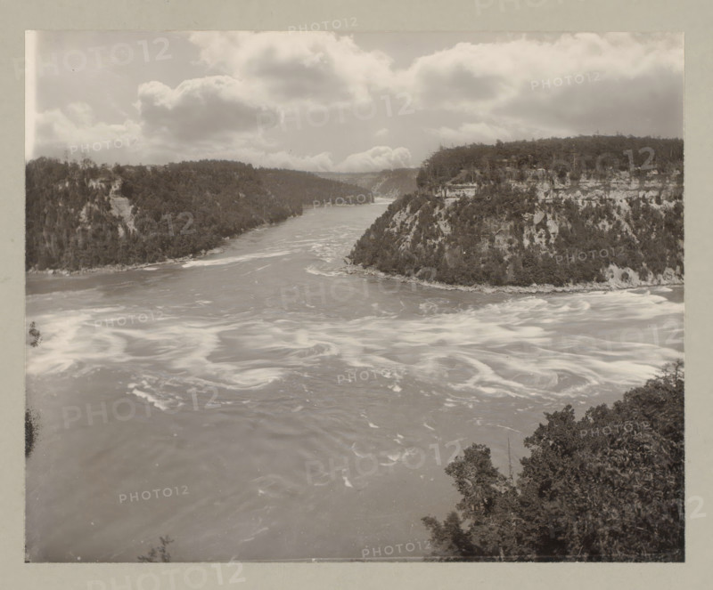 Whirlpool at Niagara Falls, United States, Canada, photograph, annotation: '11. Whirlpool.', Niagara Falls (North America), c. 1895 - c. 1915, photographer, North America, paper, pasteboard, gelatin silver print, developing gelatin silver print, height 192 mm x width 240 mm, height 243 mm x width 329 mm, A wide river flows between steep forested cliffs with swirling lighter patches on the water surface and rocky shorelines, dense trees covering the slopes and tops of the cliffs, a partly cloudy sky with large billowy clouds above, and foliage visible in the lower foreground along the river edge.