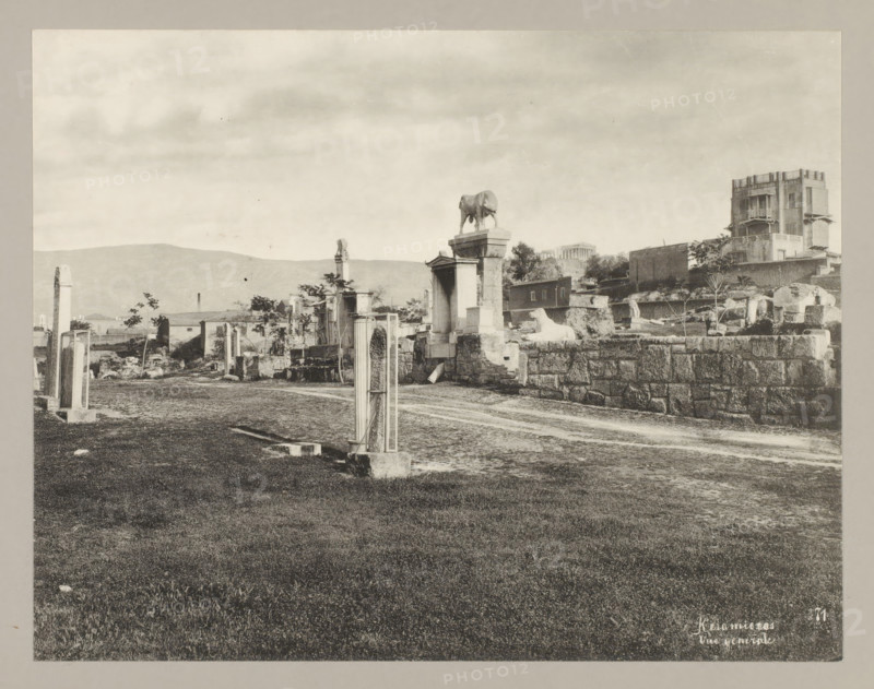 Keramiecos Vue generale 71, View of Kerameikos, photograph, annotation: '31. Cemetery at Athens.', Athens, c. 1895 - c. 1915, photographer, Athene, Periféreia Protevoúsis, pasteboard, paper, collotype, photoprint, height 215 mm x width 276 mm, height 239 mm x width 328 mm, A wide, monochrome scene showing an open grassy foreground leading to a dirt path and a ruined stone area with scattered columns and blocks. Along the path are upright stone posts and rectangular framed structures, and further back are larger stone pedestals and sculpted forms including a statue of an animal standing on a plinth and a reclining animal figure on a raised stone platform. Behind these ruins are low buildings, partial walls, and a taller multi-story building with balconies and decorative openings. In the distant background low hills are visible beneath a cloudy sky.