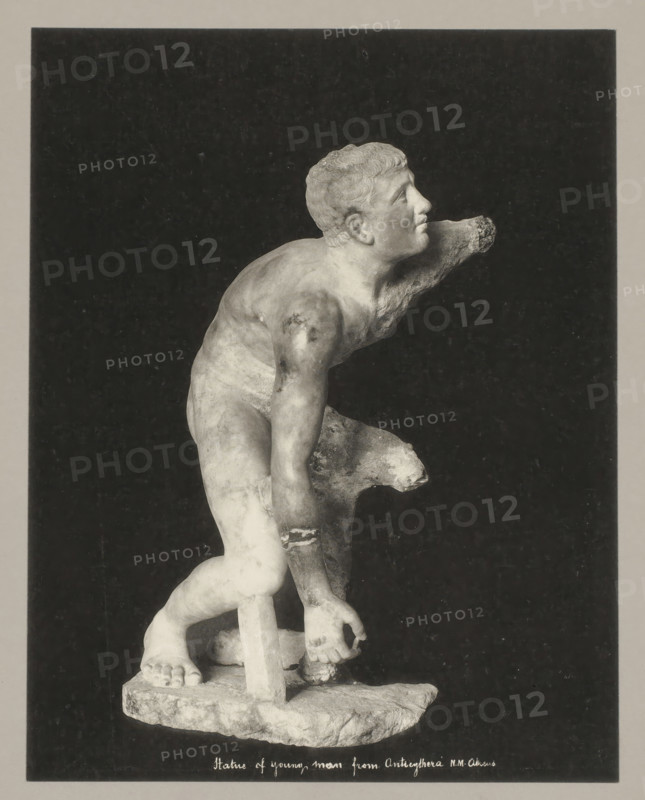 Statue of young man from Anticythera NM Athens, Statue of young man from Antikythera, photograph, number: '34.', Athens, c. 1895 - c. 1915, photographer, Athene, Periféreia Protevoúsis, pasteboard, paper, collotype, photoprint, height 274 mm x width 215 mm, height 328 mm x width 239 mm, A grayscale photograph of a partially damaged classical marble statue of a young male figure set against a dark rectangular background within a lighter border, showing the figure leaning forward with one arm extended and the other bent, a supportive tree-stump-like element behind one leg, detailed musculature and curly hair on the head turned upward, visible feet standing on a rough rectangular base, and areas of surface wear and missing fragments on the arms and torso.