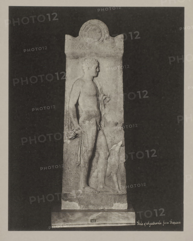 Stèle of Thepiae, Stele of Agathocles from Thepiae, photograph, number: '38.', Athens (possibly), c. 1895 - c. 1915, photographer, Athene, Periféreia Protevoúsis, pasteboard, paper, collotype, photoprint, height 273 mm x width 273 mm, height 328 mm x width 239 mm, A tall rectangular stone relief stands against a dark rectangular backdrop, showing a carved male figure in profile facing right with short curly hair and a draped garment hanging from one shoulder and arm, the figure's bare torso and legs visible and one hand holding a small object while the other hand rests near the hip, a dog-like animal stands beside the figure with its head raised, the relief has a rounded decorative top and rests on a plinth with a small rectangular plaque, the surface of the stone shows weathered texture and worn edges.