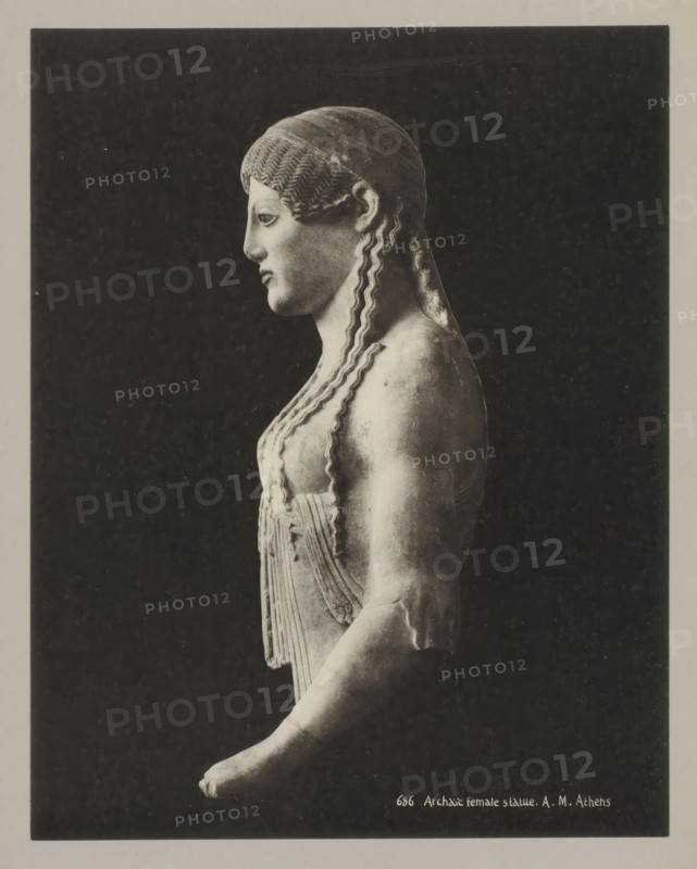 686 Archaic female statue. AM Athens, Archaic statue of a woman, photograph, number: '39.', Athens, c. 1895 - c. 1915, photographer, Athene, Periféreia Protevoúsis, pasteboard, paper, collotype, photoprint, height 274 mm x width 215 mm, height 328 mm x width 239 mm, A grayscale photograph shows a side view of a classical female statue against a dark rectangular background framed by a lighter border, depicting the statue from head to forearm; the figure has stylized wavy hair pulled back and falling in long undulating locks, a smooth profile with a straight nose and closed lips, a draped garment with vertical folds and decorative banding over the chest, a rounded shoulder and upper arm with some surface irregularities, and an extended forearm ending in a partially visible hand.