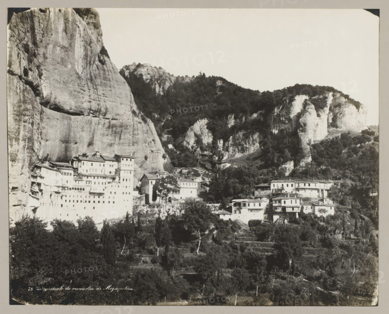 78 Vue générale du monastère de Megaspiléon, View of the monastery of Megaspileon, photograph, number: '48.', unknown, c. 1895 - c. 1915, photographer, pasteboard, paper, gelatin silver print, developing gelatin silver print, height 224 mm x width 278 mm, height 239 mm x width 328 mm, Thessaly, A black and white photograph showing a steep rocky cliff face on the left and a rugged hillside with rocky outcrops and dense tree cover occupying the center and right, with multi-story buildings of varying sizes and shapes built along the lower slopes and at the base of the cliffs, some structures appearing terraced into the rock; the foreground contains darker trees and vegetation, and the upper portion of the image shows an expanse of pale sky.