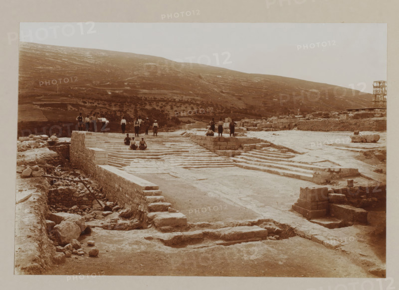 Palace of Knossos, photograph, annotation: '50. Crete, palace of Cnossos.', unknown, c. 1895 - c. 1915, photographer, pasteboard, paper, daylight gelatin silver print, height 155 mm x width 226 mm, height 239 mm x width 328 mm, Crete, Knossós, A wide sepia-toned scene of an archaeological site with broad stone terraces and stepped platforms leading up to a higher level where a group of people stand and sit; scattered stone blocks and rubble lie in the foreground and along a sunken trench with wooden beams, while low stone walls and roughly hewn blocks form retaining structures across the site; a hillside with sparse vegetation rises in the background under a pale sky, and a wooden scaffold-like structure stands near the right edge of the image.