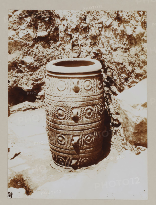 Large vase (?) at the palace of Knossos, photograph, annotation: '54. Crete. Palace at Cnossos.', unknown, c. 1895 - c. 1915, photographer, pasteboard, paper, daylight gelatin silver print, height 219 mm x width 162 mm, height 328 mm x width 239 mm, Crete, Knossós, A tall cylindrical ceramic vessel sits upright on a stone surface against a rough rock wall, the vessel featuring multiple horizontal bands of decorative raised patterns including circular rosettes, beaded rows, and twisted rope-like motifs, with protruding rounded knobs at intervals and a wide rounded rim at the top; the surrounding area shows uneven rocky textures and light and shadow contrasts on the ground and wall.