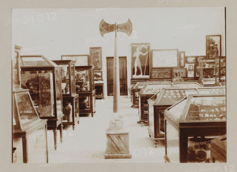 Double axe in museum on Crete, photograph, Double axe, symbol of divinity, amidst cabinets with objects., annotation: '57. Crete. Museum at Candia Double axe symbol of divinity', unknown, c. 1895 - c. 1915, photographer, pasteboard, paper, daylight gelatin silver print, height 166 mm x width 228 mm, height 239 mm x width 328 mm, A large indoor space filled with glass display cases and framed artworks is shown, with a tall vertical pole topped by a double-bladed axe positioned centrally and mounted on a square plinth at floor level, wooden-framed glass cases arranged in rows on either side containing various objects and pottery, framed paintings and panels leaning against the walls and stacked on furniture, one prominent framed image of a standing figure near the center right, wooden cabinets and pedestals supporting many of the displays, and a pale wall forming the backdrop.