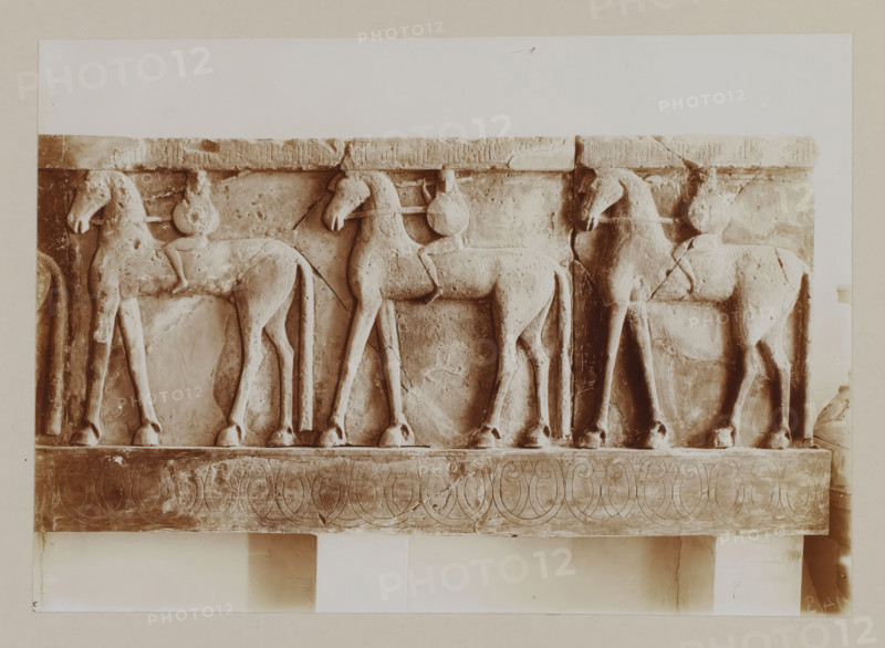 Frisian with warriors on horses, in a museum in Crete, photograph, At the bottom right the number is mirrored: 143., annotation: '62. Muzeum in Candia', unknown, c. 1895 - c. 1915, photographer, pasteboard, paper, daylight gelatin silver print, height 167 mm x width 233 mm, height 239 mm x width 328 mm, A carved stone frieze depicting a row of standing horses in profile with simple, rounded bodies and elongated legs, each horse shown with a bridle and a circular ornament near its neck, mounted on a rectangular panel with a patterned lower band of interlocking curved motifs, the surface showing cracks, chips, and weathered texture, and the entire relief presented against a pale background with visible borders.