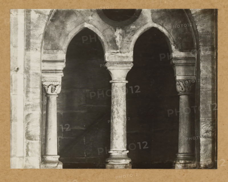 Eastern choir, inner window, View of a window of Bamberg Cathedral, album page, photograph, This photo is part of an album., number: '10', Bamberg, 1900 - c. 1920, photographer, Bavaria, paper, baryta paper, gelatin silver print, developed gelatin silver print, height 168 mm x width 217 mm, church (exterior), Bamberg Cathedral, A close view of a stone architectural opening featuring three arched openings supported by rounded columns with molded capitals and bases, the stone showing weathered textures and uneven coloration, decorative carved leaf motifs on the outer capitals, a dark recessed background visible through the arches, and surrounding vertical stone jambs with worn surfaces.