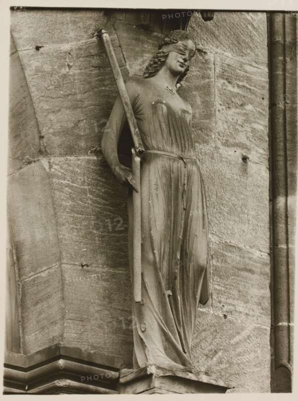 Detail of the Fürstenportal in Bamberg Cathedral, Fürstentor, Synagogue v. halblinks, album page, photograph, This photo is part of an album., inscription: 'Kunsthist. Seminar, Marburg (Lahn), Photogr. Abteilung, Vervielfältigung, untersagt.', number: '103', Bamberg, 1900 - c. 1920, photographer, Bavaria, paper, baryta paper, gelatin silver print, developing gelatin silver print, height 239 mm x width 180 mm, church (exterior), Bamberg Cathedral, A stone statue of a robed, blindfolded female figure is shown in close-up against a textured stone wall, the figure standing with a long staff held in one hand and the other hand resting near her hip, her draped garment falling in vertical folds to a carved base, her wavy hair framed by the blindfold and her head tilted slightly, with visible weathering and small spots on the statue and surrounding masonry.