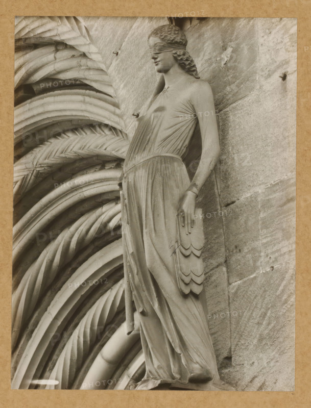 Detail of the Fürstenportal in Bamberg Cathedral, Fürstentor, Synagogue v. Halbrechts. 106a, dasselbe, album page, photograph, This photo is part of an album., number: '106.', Bamberg, 1900 - c. 1920, photographer, Bavaria, paper, baryta paper, gelatin silver print, developing gelatin silver print, height 211 mm x width 160 mm, church (exterior), Bamberg Cathedral, A grayscale photograph of a sculpted female figure set against a textured stone background, the figure wearing a long draped gown with visible folds and a belted waist, a blindfold covering the eyes, wavy hair pulled back, one arm hanging by the side with a layered decorative element resembling feathers, and the other arm partly visible; to the left of the figure a series of rounded, ribbed, and braided architectural carvings form a repeating pattern that fills the background.