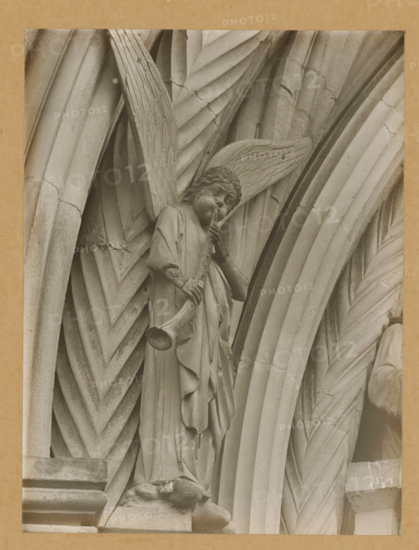 Detail of the Princely Portal in Bamberg Cathedral, Princely Gate, Synagogue, Posaunenengel vl, album page, photograph, This photo is part of an album., number: '107.', Bamberg, 1900 - c. 1920, photographer, Bavaria, paper, baryta paper, gelatin silver print, gelatin silver developing print, height 217 mm x width 163 mm, church (exterior), Bamberg Cathedral, A stone carving of an angel with textured wings stands within a series of arched, ribbed stone surfaces; the angel wears flowing robes with visible folds and holds a long trumpet to its lips while one hand supports the instrument, the face showing carved features and wavy hair; surrounding the figure are layered, chevron-patterned and fluted stone arches with weathered surfaces and small chips and cracks, all shown in a monochrome tone against a plain border.