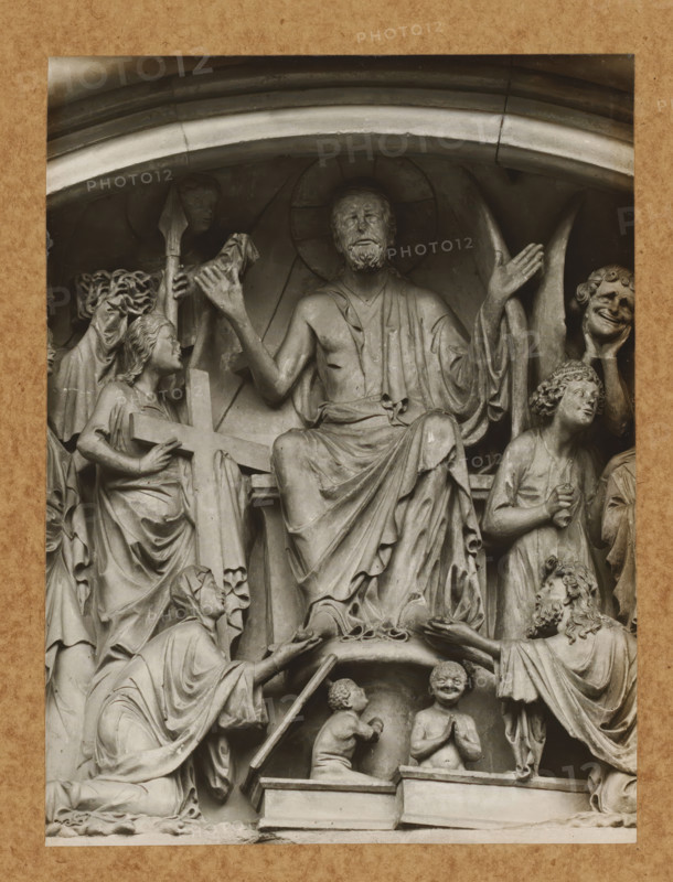 Detail of the tympanum of the Fürstenportal in Bamberg Cathedral, Fürstentor, Tymphanon, Christ from the front, album page, photograph, This photo is part of an album., number: '113', Bamberg, 1900 - c. 1920, photographer, Bavaria, paper, baryta paper, gelatin silver print, height 217 mm x width 163 mm, church (exterior), Bamberg Cathedral, A carved stone relief showing a seated bearded man with a halo and draped robes, his arms raised and palms open, surrounded by multiple figures in flowing garments; to his left a kneeling woman holds a large cross while another figure reaches upward with a spear-like object, to his right a standing figure with curly hair looks upward and a smiling masked face is held by a hand; below the central figure two small childlike figures kneel on a platform, one with hands together and the other turned toward the central figure, while a robed figure on the lower left kneels and extends an object toward the central scene.