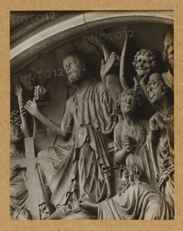 Fürstentor, Tymphanon, Christ the Lady, Detail of the tympanum of the Fürstenportal in Bamberg Cathedral, album page, photograph, This photo is part of an album., number: '115', Bamberg, 1900 - c. 1920, photographer, Bavaria, paper, baryta paper, gelatin silver print, gelatin silver developing print, height 208 mm x width 163 mm, church (exterior), Bamberg Cathedral, A close-up photograph of a carved stone relief framed by a textured border, showing multiple sculpted figures in draped robes and detailed hair. One central seated figure has a beard and raised arm with an open hand, while surrounding figures display varied facial expressions and gestures, including a smiling figure with stylized curls and a youthful figure looking upward. Folded and flowing drapery covers limbs and torsos, and carved architectural arches appear above the group. A partial cross is visible and several hands are clasped or holding small objects, with a smaller figure kneeling at the lower edge. The stone sur...