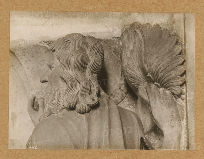 Georgenchorschranken 3 P. vr (Profil), Detail of the rood screen in the East Choir of Bamberg Cathedral, album page, photograph, This photo is part of an album., number: '126', Bamberg, 1900 - c. 1920, photographer, Bavaria, paper, baryta paper, gelatin silver print, developed gelatin silver print, height 167 mm x width 225 mm, interior of church, Bamberg Cathedral, A close-up photograph of a carved stone figure shown from the shoulders up and turned in profile, featuring wavy hair and a full beard with carved curls, a pronounced nose, and a visible ear; the figure wears a draped garment over the shoulders and holds one hand near the chest; next to the figure is a carved wing with layered feathers and a hand positioned beneath the wing; the stone surface shows variations in texture and subtle weathering, and the scene is bordered by a rectangular frame with a textured outer mat.