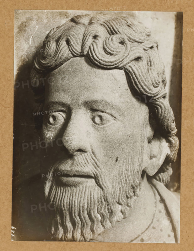 Georgenchorschranken 4 Avl (Heads from front), Detail of the rood screen in the East Choir of Bamberg Cathedral, album page, photograph, This photo is part of an album., number: '129', Bamberg, 1900 - c. 1920, photographer, Bavaria, paper, baryta paper, gelatin silver print, developed gelatin silver print, height 227 mm x width 165 mm, interior of church, Bamberg Cathedral, A close-up black and white photograph of a carved stone head mounted on a textured background, showing the upper face, hair, and beard. The stone figure has stylized wavy hair with carved concentric curls on the top and sides, deeply incised lines in the beard forming vertical ridges, a pronounced mustache, and a smooth forehead. The eyes are rounded with carved pupils and surrounding eyelids, and the nose is broad with subtle weathering marks. The surface of the stone shows variations in tone and small pitted spots, with darker shadowed areas along the left side of the face and under the chin. The photograph itself...