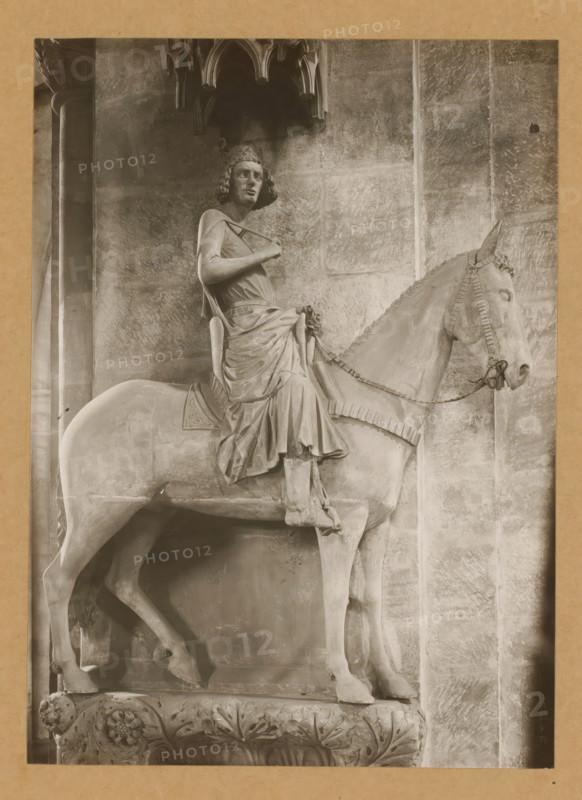 Bamberger Reiter in the cathedral of Bamberg, Der Reiter, Knitting page, album page, photograph, This photo is part of an album., number: '131.', Bamberg, 1900 - c. 1920, photographer, Bavaria, paper, baryta paper, gelatin silver print, developed gelatin silver print, height 222 mm x width 160 mm, riding a horse, ass, or mule, rider, horseman, interior of church, Cathedral of Bamberg, A stone equestrian statue occupying most of the frame shows a robed rider seated on a bridled horse, both carved with detailed drapery and tack; the rider wears a crown-like headpiece and has curly hair, a cloak draped over one shoulder, and one hand holding a strap across the chest while the other rests near the waist, the folds of the robe falling over the horse's flank and legs; the horse stands with a raised head, visible bridle and bit, and textured mane, its legs positioned on a carved rectangular plinth decorated with acanthus leaves and floral motifs; the background consists of a stone wall with v...