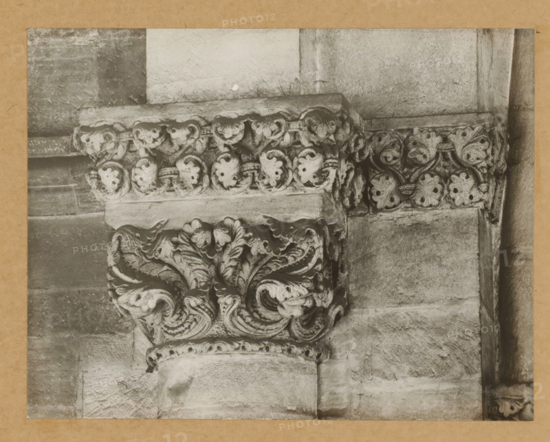 East church, with Triumphal arch chapter, Capital of Bamberg Cathedral, album page, photograph, This photo is part of an album., number: '13.', Bamberg, 1900 - c. 1920, photographer, Bavaria, paper, baryta paper, gelatin silver print, height 165 mm x width 214 mm, church (exterior), Bamberg Cathedral, A close-up view of a carved stone capital and surrounding masonry showing ornate acanthus leaf and scroll motifs in a weathered light-toned stone, with a horizontal band of rounded carved elements above and textured blocks of stone forming the wall and column surfaces, all framed by a narrow darker border.