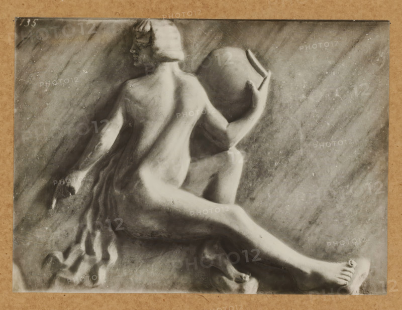 Grabmal, Prudentia, Detail of the tomb of Pope Clement II in the cathedral of Bamberg, album page, photograph, This photo is part of an album., number: '135', Bamberg, 1900 - c. 1920, photographer, Bavaria, paper, baryta paper, gelatin silver print, developing gelatin silver print, height 166 mm x width 226 mm, Person, Clemens II, interior of church, Bamberg Cathedral, A grayscale photograph of a sculpted relief showing a seated nude figure viewed from behind and in profile, with a bobbed hairstyle and turned head, one arm bent and holding a large round vessel against the shoulder while the other arm rests downward, draped fabric cascading beside the figure, a textured stone background with diagonal streaking and surface blemishes, and the image bordered by a light brown mat.