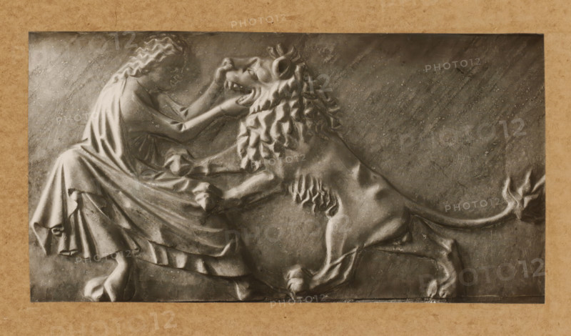 Detail of the tomb of Pope Clement II in the cathedral of Bamberg, Grabmal, Stärke, album page, photograph, This photo is part of an album., number: '140', Bamberg, 1900 - c. 1920, photographer, Bavaria, paper, baryta paper, gelatin silver print, developing gelatin silver print, height 115 mm x width 220 mm, Person, Clemens II, interior of church, Bamberg Cathedral, A rectangular relief panel set against a textured border depicts a seated person with long wavy hair and draped clothing facing a large lion; the person’s arms reach toward the lion’s head and the lion rears with a flowing mane and open mouth, its front paws raised and hind legs braced, with detailed musculature and fur textures carved across both figures and a smooth, slightly scratched background.