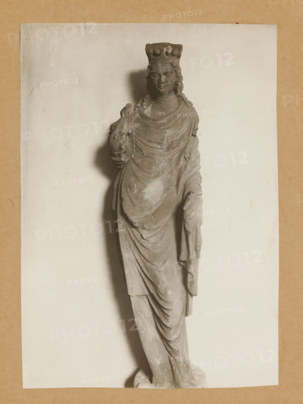 Sculpture in the cloister of Bamberg Cathedral, Cloister of St. Catherine vv, album page, photograph, This photo is part of an album., number: '143.', Bamberg, 1900 - c. 1920, photographer, Bavaria, paper, baryta paper, gelatin silver print, developed gelatin silver print, height 227 mm x width 161 mm, interior of church, Bamberg Cathedral, A grayscale photograph of a standing stone statue depicting a crowned figure draped in flowing robes, shown from head to feet, with a serene facial expression and long wavy hair; the figure holds a small animal against the chest with one hand while the other hand rests by the side, and the statue casts a soft shadow on a plain light background framed by a textured neutral border.