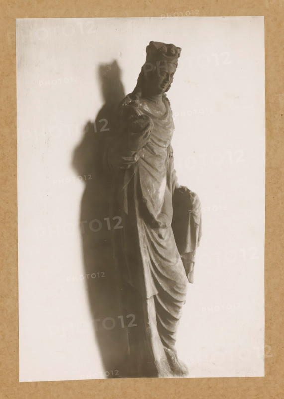 Cloister of St. Catherine of the People, Sculpture in the cloister of Bamberg Cathedral, album page, photograph, This photo is part of an album., number: '144', Bamberg, 1900 - c. 1920, photographer, Bavaria, paper, baryta paper, gelatin silver print, developed gelatin silver print, height 230 mm x width 157 mm, interior of church, Bamberg Cathedral, A grayscale photograph of a draped statue showing a standing figure wearing a crown and layered robes, the figure's head slightly tilted and one arm bent at the elbow while the other draped hand holds fabric, with a strong shadow cast to the left on a pale background and a textured border around the image.