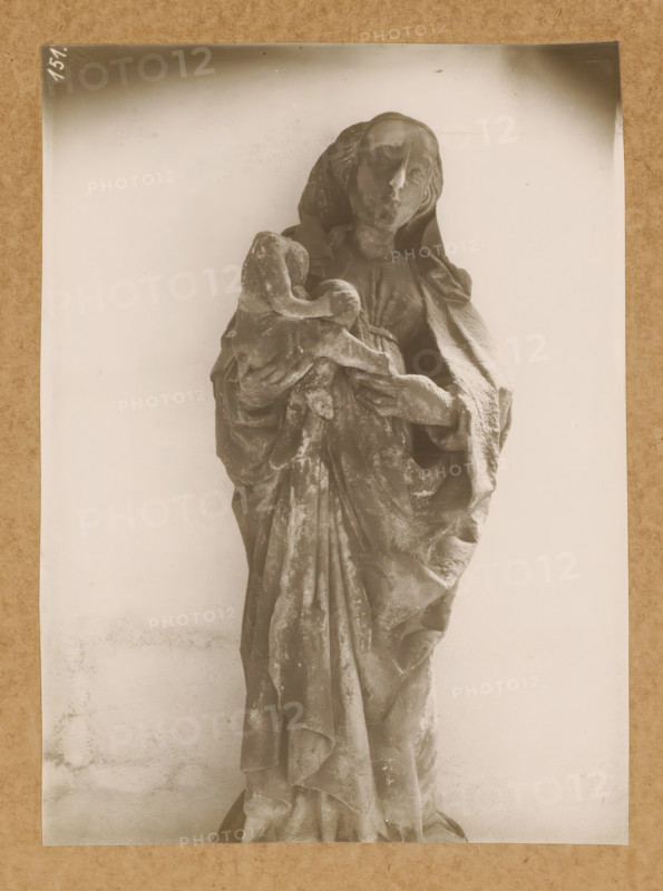 Cloister, Madonna, Sculpture in the cloister of Bamberg Cathedral, album page, photograph, This photo is part of an album., number: '151.', Bamberg, 1900 - c. 1920, photographer, Bavaria, paper, baryta paper, gelatin silver print, height 228 mm x width 168 mm, interior of church, Bamberg Cathedral, A monochrome photograph of a carved figure draped in flowing robes, shown from head to lower torso, standing against a plain light background. The figure has a serene facial expression with closed or lowered eyes and hair partially covered by a hood or veil. One arm cradles a smaller wrapped form while the other supports it, with detailed folds and texture in the garments visible. The surface of the carving shows wear and roughness, and the image is bordered by a narrow darker edge against a textured mat.
