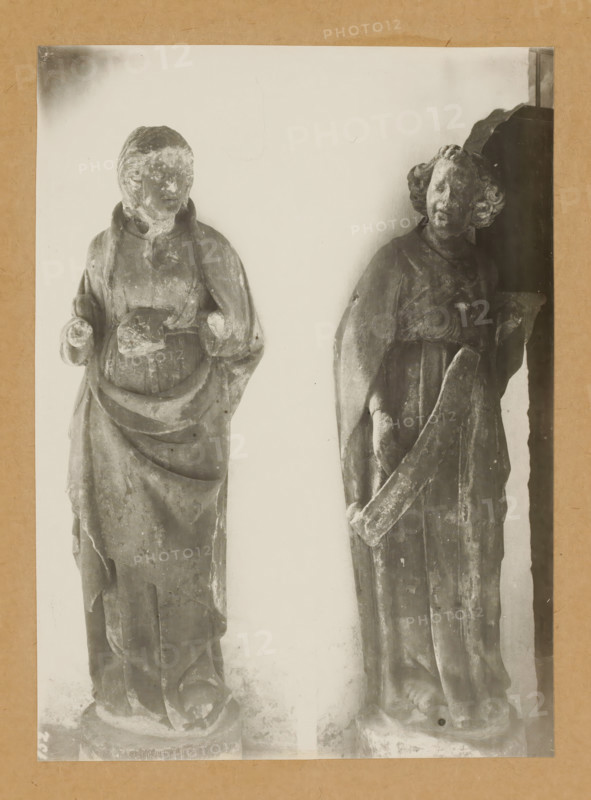 Cloister, Saint (Reverence), Sculpture in the cloister of Bamberg Cathedral, album page, photograph, This photo is part of an album., number: '152', Bamberg, 1900 - c. 1920, photographer, Bavaria, paper, baryta paper, gelatin silver print, height 225 mm x width 164 mm, interior of church, Bamberg Cathedral, Two weathered stone statues stand against a pale background, each mounted on a round base and framed by a neutral border. The statue on the left depicts a robed figure with a hooded or draped garment, hands positioned near the midsection, and a rounded, worn face with indistinct features; the robe shows layered folds and surface erosion. The statue on the right shows a robed figure with curly hair, a tilted head, and a long flowing garment; one arm is bent holding a long curved object while the other arm hangs by the side, and the surface displays chips, scratches, and areas of wear. Both statues exhibit aged textures, weathering marks, and variations in tonal contrast.
