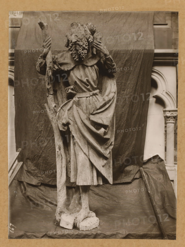 Cathedral treasure, St. Christopher from the front, Wooden statue of Christopher of Lycia in the cathedral of Bamberg, album page, photograph, This photo is part of an album., number: '167', Bamberg, 1900 - c. 1920, photographer, Bavaria, paper, baryta paper, gelatin silver print, developed gelatin silver print, height 231 mm x width 171 mm, Person, Christopher of Lycia, interior of church, Cathedral of Bamberg, A weathered stone statue of a robed standing figure is shown against a draped cloth backdrop, the figure holding a long staff in one hand and a smaller object near its shoulder with the other, the head featuring a full curly beard and worn facial details, the robe displaying deep folds and a belted waist, the lower garment hanging in layered drapery over thin legs and worn feet standing on a rough base, the scene framed by portions of architectural stonework visible at the sides and a wooden pole leaning against the draped backdrop.
