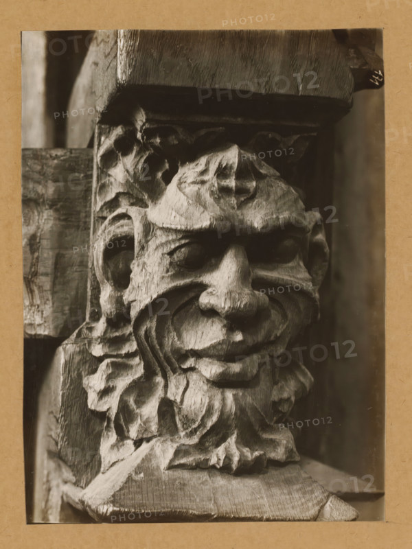 Choir stalls in the St. Peter's choir, Mask, Detail of the choir stalls in Bamberg Cathedral, album page, photograph, This photo is part of an album., number: '171', Bamberg, 1900 - c. 1920, photographer, Bavaria, paper, baryta paper, gelatin silver print, developed gelatin silver print, height 223 mm x width 164 mm, interior of church, Bamberg Cathedral, A close-up sepia-toned photograph of a carved wooden face with exaggerated features, showing deep wood grain and chisel marks; the face has prominent brows, hollowed eyes, a broad nose, pronounced cheeks, and a curled mustache or beard element that blends into surrounding carved foliage-like forms, with a rectangular wooden beam above and textured wooden surfaces flanking the carving.